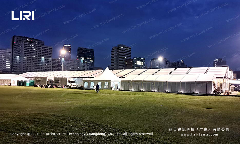 Night overview of illuminated clear-span tent on football field with city skyline