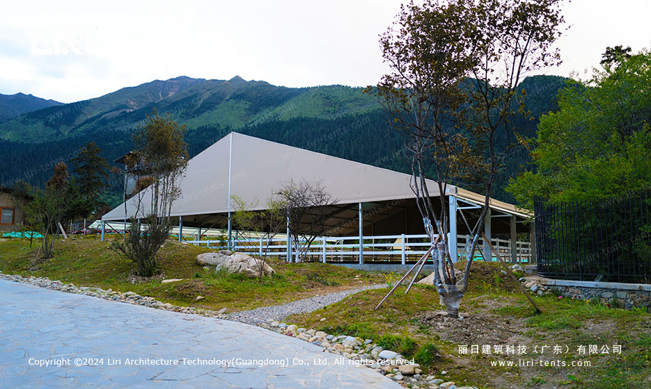Clear span marquee standing on sloping ground below a forested mountain with a gravel path and low white railing